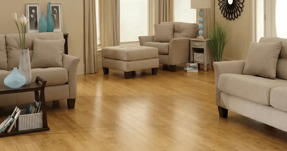 A living room with professionally installed bamboo flooring, beige sofas, a matching ottoman, a side table with a blue lamp, vases, framed pictures, and large windows with blinds and curtains in Houston, TX