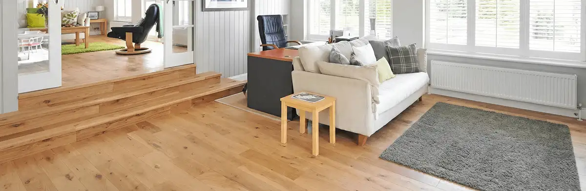 A modern living room with professionally installed laminate flooring, a beige sofa, a small side table, a gray rug, and large windows with white shutters in Houston, TX
